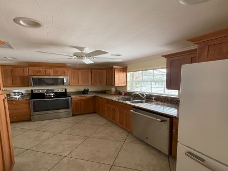 A bright kitchen with wooden cabinets, granite counters, a stainless stove and sink, tiled floor, and ceiling fan.