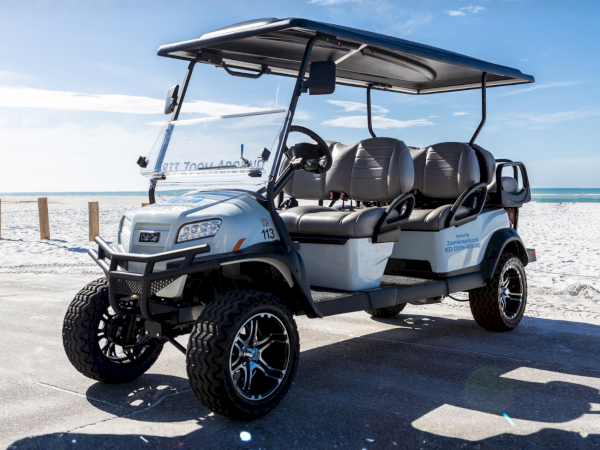 A customized four-seat Club Car golf cart on a sunny seaside boardwalk, with sand, ocean, and a blue sky in the background.
