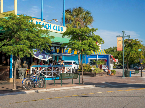 Outdoor Beach Club with a &ldquo;Beach Club&rdquo; sign, diners under awnings, bikes parked outside, and palm trees along the street.