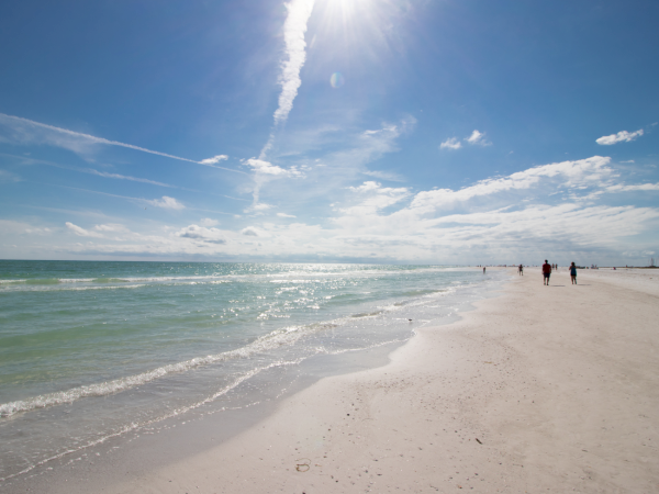 A sunny sandy beach with waves rolling in and people walking, under a blue sky with a long jet contrail.