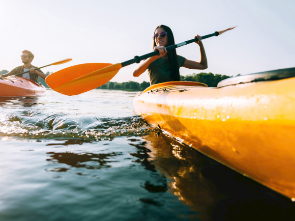 Two people paddle orange kayaks on a calm lake, with a woman in front smiling and steering with her paddle.