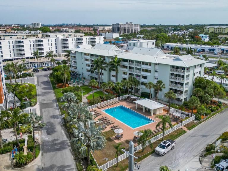 Aerial view of a residential complex with white apartment blocks, a blue swimming pool, palm trees, roads, and parked cars.