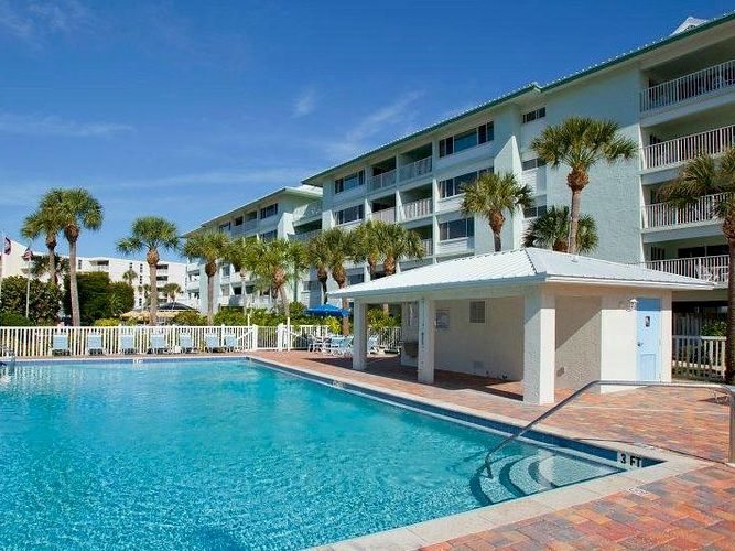 A sunny hotel courtyard with a blue swimming pool, white buildings, and palm trees.