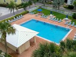An aerial view of a hotel with a large blue swimming pool, surrounded by sun loungers and palm trees.