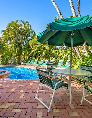A sunny resort pool with a green umbrella, metal loungers, and lush palm gardens.