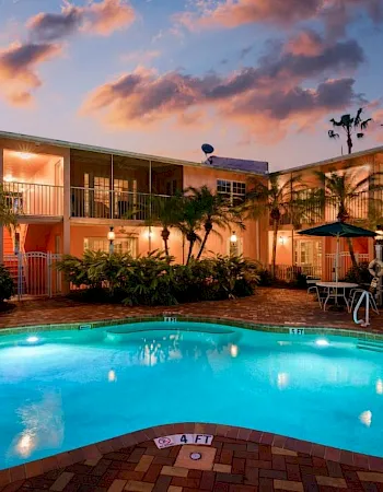 A hotel courtyard at sunset with a bright swimming pool, palm trees, and lounge chairs in front of two-story rooms.