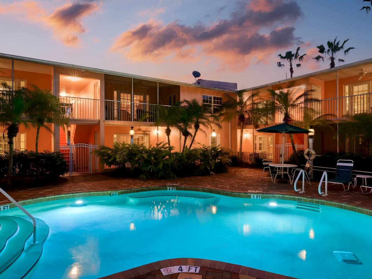 A resort courtyard at sunset with a swimming pool, patio lights, palms, and lounge chairs in front of hotel rooms.