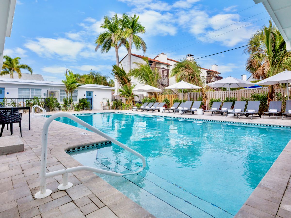 Sunny backyard pool with blue water, lounge chairs under white umbrellas, palm trees, and white buildings under a clear sky.