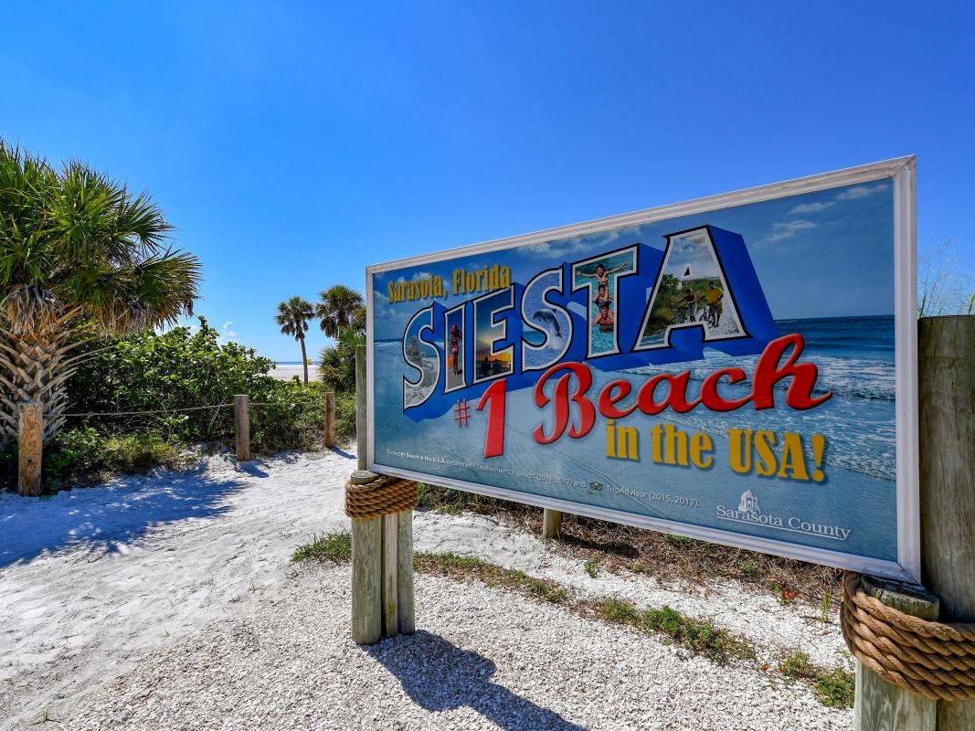 A sign reading &ldquo;Siesta Key Beach in the USA!&rdquo; with &ldquo;Sarasota, Florida&rdquo; on a sunny beach path.