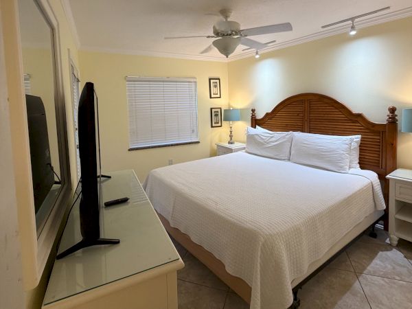 A bright bedroom with a wooden bed, ceiling fan, window blinds, nightstand lamps, and a wall TV/mirror.