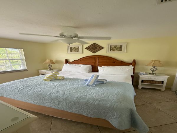 A bright bedroom with a wooden bed, white linens, bedside lamps, wall art, and a ceiling fan overhead.